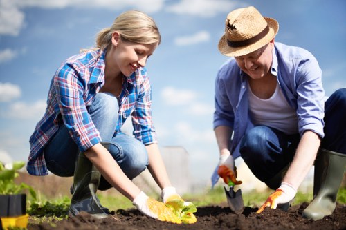 In a bright outdoor garden setting, a young woman and an older woman are engaged in planting activities on a clear, sunny day. The younger woman, dressed in a blue checkered shirt and jeans, is kneeling on the soil, handling a yellow gardening glove and tending a small plant or seedling in freshly turned soil, which appears rich and dark. Beside her, the older woman wears a straw sunhat, a light jacket over a white top, and jeans, and is also kneeling while using a small garden trowel to work the soil carefully. The garden features neatly maintained flower beds with low, green foliage and possibly small flowering plants, bordered by paving slabs or wooden edging. In the background, there are hints of other garden features, such as lawn areas and possibly more plants, under a blue sky with some clouds, indicating good weather conditions. The scene reflects active gardening and outdoor maintenance suitable for a professional landscaping and gardening service website focused on enhancing local outdoor spaces in Lewisham or nearby areas.