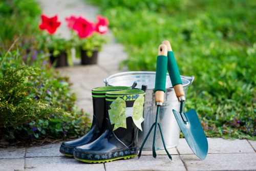 A collection of gardening tools and supplies arranged outdoors on a paved surface in a garden setting. In the foreground, there are a pair of black rubber gardening boots with a decorative pattern, with a yellow and green patterned towel draped over one of them. Next to the boots, a metal bucket contains a pair of hand trowels with green handles, a garden fork, and a small gardening spade, all with wooden handles. Behind the tools, a white plastic container or watering can is partially visible. The background features lush green foliage, flowering plants with bright pink blooms in pots, and a stone pathway extending into the garden. The scene suggests preparations for outdoor gardening or planting activities in a well-maintained garden environment, possibly in Lewisham, with natural daylight and a hint of a cloudy sky, emphasizing the typical conditions for garden work during spring or summer months.