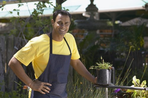A smiling man dressed in a yellow T-shirt and dark apron stands in a well-maintained garden, holding a small black plastic pot with a young, green plant. The garden features lush grass, a variety of shrubs, and a flower bed with blooming plants. In the background, there are trees, a wooden fence, and garden structures, indicating a landscaped outdoor space typical of residential gardens in Lewisham. The scene suggests outdoor gardening or planting activity performed in a bright, clear weather condition, with natural light illuminating the area. The setting highlights typical elements of professional gardening services, such as plant care, planting, and garden maintenance, relevant to residents seeking expert landscaping or gardening support from companies like Landscapers Lewisham.