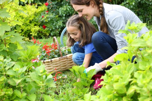 A woman and a young girl are engaged in gardening in a lush, well-maintained backyard. The woman, with long brown hair, is kneeling and smiling as she helps the girl pick flowers from a flower bed with vibrant red blossoms and green foliage. The girl, wearing a blue top, is holding a wicker basket with a handle, partially filled with flowers or garden produce. In the background, there are neatly trimmed hedges and a variety of plants, suggesting a thoughtfully landscaped outdoor space. The ground is covered with a mixture of grass and soil, with some areas showing fresh earth. The overall scene is bathed in natural daylight, emphasizing the healthy greenery and bright colours of the garden. This peaceful outdoor environment demonstrates typical garden elements that a professional gardening service like Landscapers Lewisham might maintain or enhance, reflecting a tidy and flourishing yard in the Lewisham area, with a focus on cultivating a welcoming and vibrant garden space.