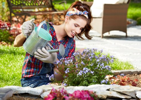 A young woman with brown hair tied back and wearing a blue and red plaid shirt, a colorful headband, and gardening gloves, is kneeling in a landscaped garden in Lewisham. She is watering a small flower bed filled with purple and pink flowering plants using a metal watering can. The garden features a neatly edged flower bed with dark soil, surrounded by a low stone border. In the background, there is a well-maintained lawn with lush green grass, a wooden deck or patio area, and outdoor furniture, suggesting a tidy and inviting outdoor space. The scene is set in bright daylight with natural sunlight, highlighting the vibrant colors of the flowers and greenery, and indicating good weather suitable for gardening activities. This setting highlights outdoor maintenance and gardening services, particularly flower bed creation and plant care, offered by Landscapers Lewisham in the local area.