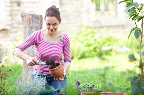 A young woman with dark hair pulled back, wearing a pink and white striped long-sleeve top, is engaged in gardening activities in a well-maintained backyard garden. She is holding a clay flower pot with dark soil inside, using a small trowel to scoop or spread the soil. Surrounding her are garden elements including a lush, green lawn, flower beds with purple and green foliage, and potted plants. In the background, there are trees, a stone or brick wall, and a weathered tree stump, indicating a landscaped outdoor space typical of residential gardens in Lewisham. The garden has a natural, inviting appearance, with fresh and healthy plants, and is bathed in natural daylight, suggesting a clear weather day. This scene exemplifies garden maintenance and planting activities that landscapers Lewisham may provide as part of their comprehensive gardening services.
