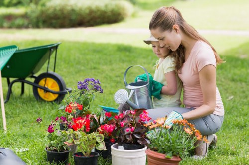 In a well-maintained garden with lush green grass, a woman and a young girl are engaged in gardening activities near a colorful array of potted flowers and plants, including vibrant red, pink, purple, and orange blooms. The garden features a neatly mowed lawn, with a garden wheelbarrow positioned to the left, indicating ongoing outdoor maintenance. Behind them, there are mature trees and a hedge line providing privacy and natural border. The weather appears sunny and bright, casting natural light over the scene, suitable for outdoor gardening. The woman is guiding the girl, both wearing casual clothing and gardening gloves, as they tend to the flower pots, suggesting a family-focused outdoor space. The setting reflects a typical landscaped garden in Lewisham, often managed or improved by local gardening services from Landscapers Lewisham, who specialize in lawn care, plant maintenance, and outdoor aesthetic enhancements.