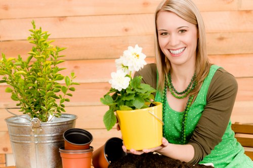 A well-maintained back garden in Lewisham featuring a lush, dense green lawn with uniform grass height, bordered by flower beds and shrub borders. In the foreground, a young woman in a green polka-dot apron is smiling while holding a yellow flower pot with a flowering plant, surrounded by gardening tools and empty plant pots. Behind her, a wooden fence provides privacy, and various garden elements such as a small potted evergreen shrub in a metal bucket and a wooden garden bench are visible. The outdoor space appears to be on a clear, sunny day with natural light illuminating the scene, highlighting the vibrant greens of the lawn and plants, and the natural textures of paving and soil areas. The garden reflects careful landscaping and planting, aligned with professional gardening services such as those offered by Landscapers Lewisham, in a residential area within the Lewisham postcode, emphasizing a neat and inviting outdoor environment suitable for outdoor activities and relaxation.