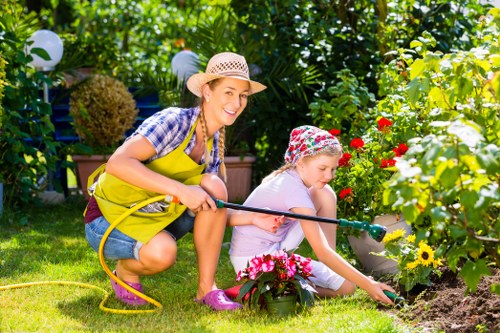 A woman and a young girl gardening together in a lush backyard garden, with the woman kneeling on the grass holding a garden hose and smiling at the camera, while the girl is focused on planting or watering flowers in a flower bed bordered by vibrant, multi-colored blooms including yellow and red flowers. The garden features a well-maintained lawn, dense green foliage, and garden ornaments such as white spherical lights strung along a hedge or trellis in the background. Sunlight filters through the leafy canopy, illuminating the scene with natural light, indicative of a bright, clear day. The setting suggests a family-friendly outdoor space suitable for gardening, lawn care, and landscaping activities, with ample space for plant growth and aesthetic landscaping elements. This scene reflects outdoor garden maintenance and planting activities typical of residential gardens serviced by local landscapers like Lewisham residents from Landscapers Lewisham, emphasizing a vibrant, healthy garden environment in a suburban setting.