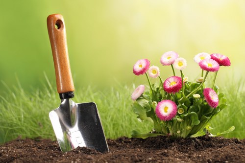 A close-up view of a garden scene in a backyard or front garden, showing a small flower bed with a cluster of pink and white daisy-like flowers growing amidst lush green foliage. The soil surrounding the plants appears freshly turned, suggesting recent planting or soil tending. In the foreground, a garden trowel with a wooden handle and metal blade is partially embedded in the dark, rich soil. The background features a softly blurred green lawn and possibly some hint of garden foliage, indicating a well-maintained outdoor space suitable for gardening services in Lewisham or surrounding areas. The natural daylight and mild weather conditions create soft lighting, emphasizing the vivid colors of the flowers against the vibrant green backdrop, aligning with garden maintenance, planting, and landscaping activities common to local gardens managed by companies like Landscapers Lewisham.